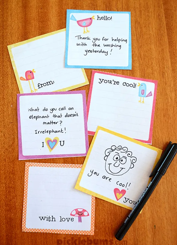 Square lunch box notes are seen on a wooden table with a black pen. These free printable lunch box notes are blank and can be customized to your child or spouse. They all have a colored border and small illustrations of birds, hearts, and mushrooms. These drawings are accompanied by small phrases like