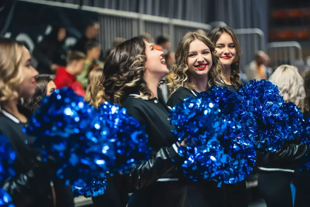 Energetic cheerleaders in uniform, with pom-poms, hyping up the crowd at a basketball game in a brightly lit arena, creating an exciting atmosphere for players and fans.