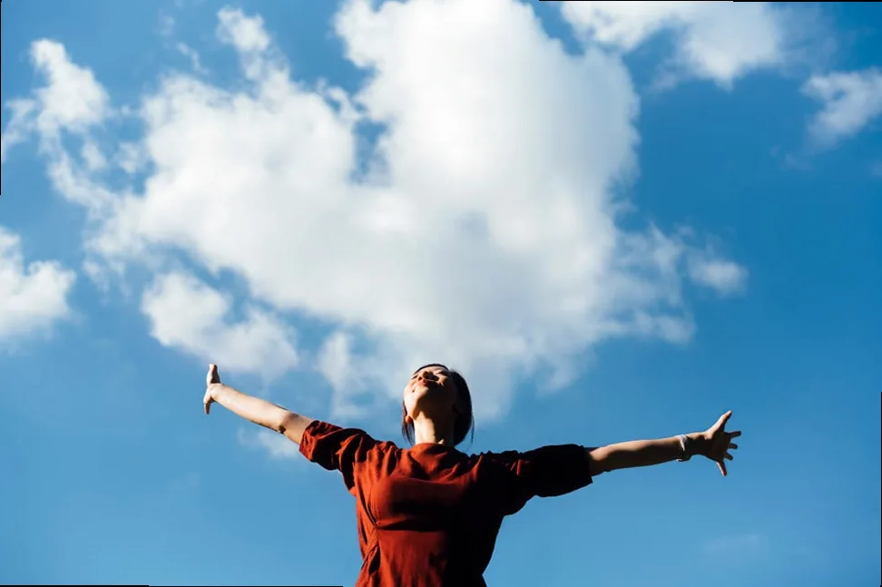 Low angle portrait of a young Asian woman with her eyes closed, stretching arms upwards, embodying freedom and relief against a blue sky, symbolizing self-love and connection with nature.
