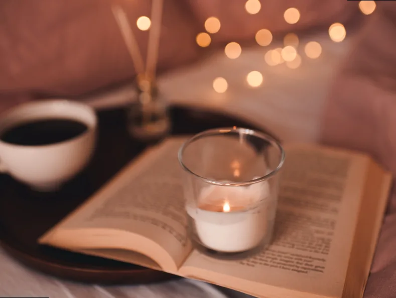 Warm, ambient lighting with a lit candle and a cup of tea next to an open book, setting a cozy reading mood.