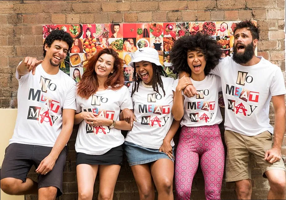 Five people stand in front of an exposed brick wall, smiling and laughing wearing No Meat May tshirts.