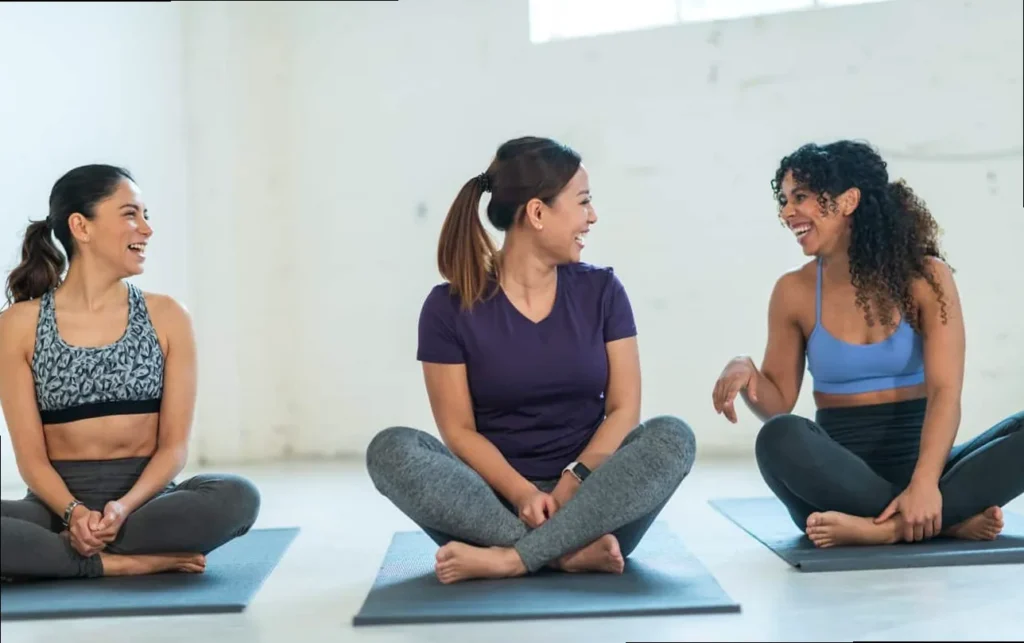two friends sitting on yoga mats laughing, embracing the joy of a shared, lighthearted yoga practice