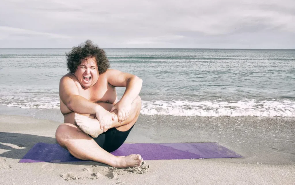 a man laughing joyfully while doing yoga on a sunny beach, embodying relaxed and goofy enjoyment