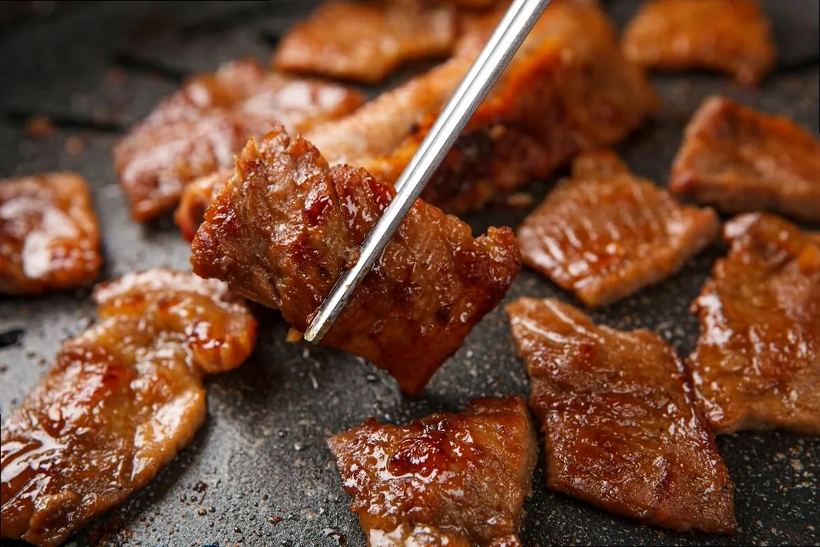 Close-up of sizzling Korean barbecue meat being cooked on a tabletop grill with chopsticks.