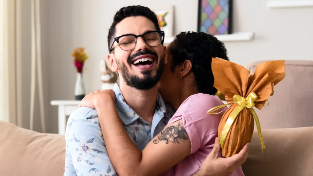 A joyful couple embraces on a sofa. The man, wearing glasses and a floral shirt, smiles. The woman, in a pink shirt, holds a wrapped gift.