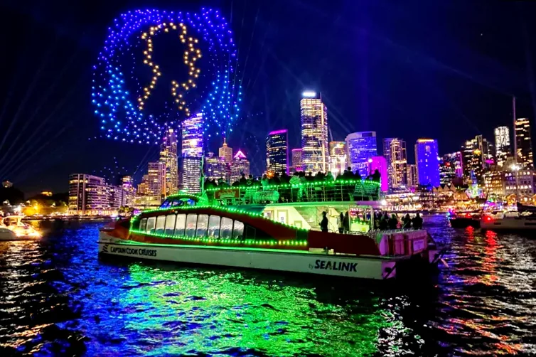 A catamaran ferry on Sydney Harbour at night, with the city lights and Vivid installations in the distance.