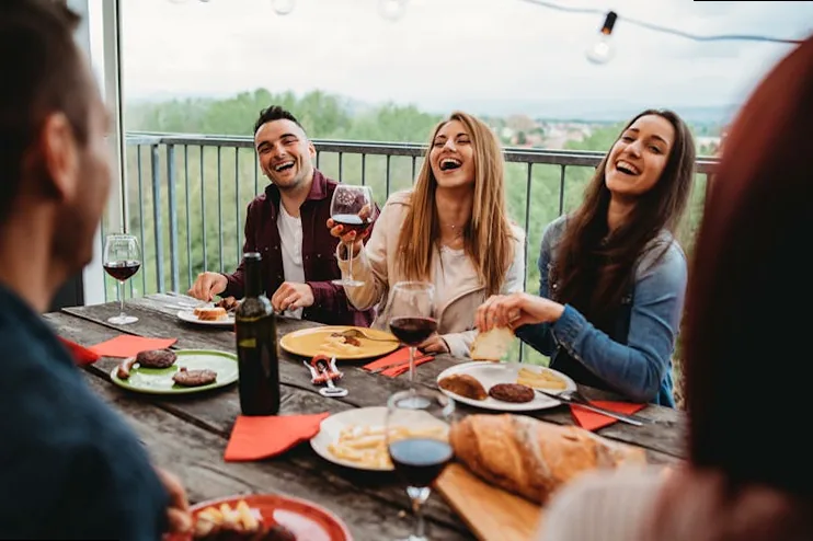 Diverse group of friends laughing and enjoying a Friendsgiving meal with food and drinks on a table, showcasing candid celebration and chosen family.