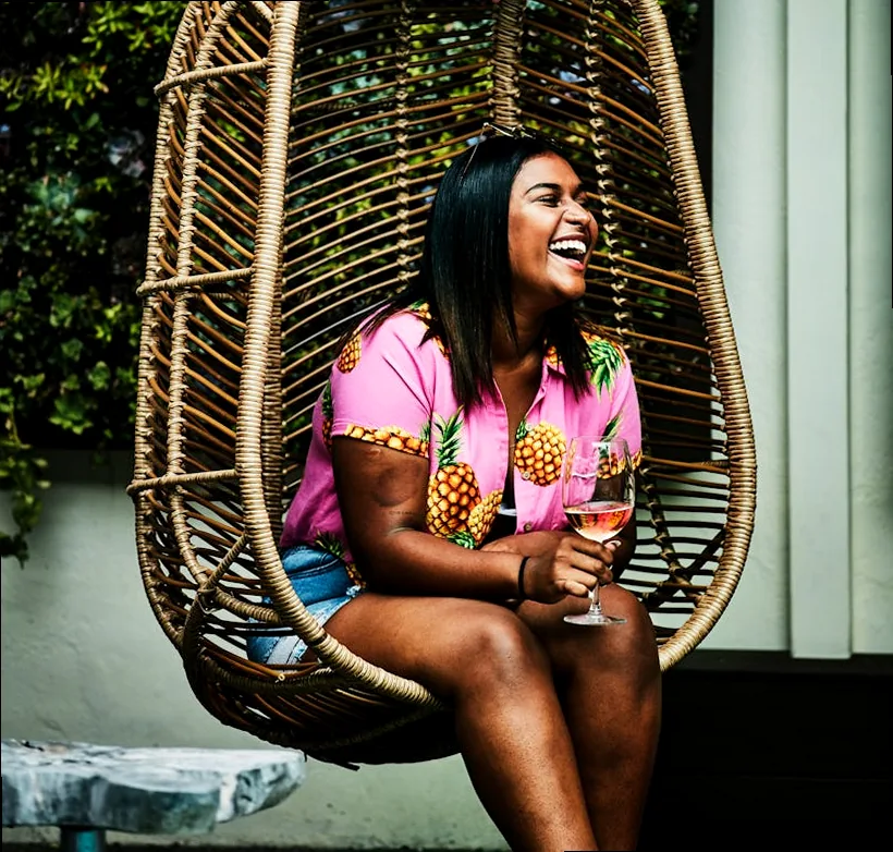 Young woman sips rosé, laughing in a chic hanging chair. Perfect summer vibe.