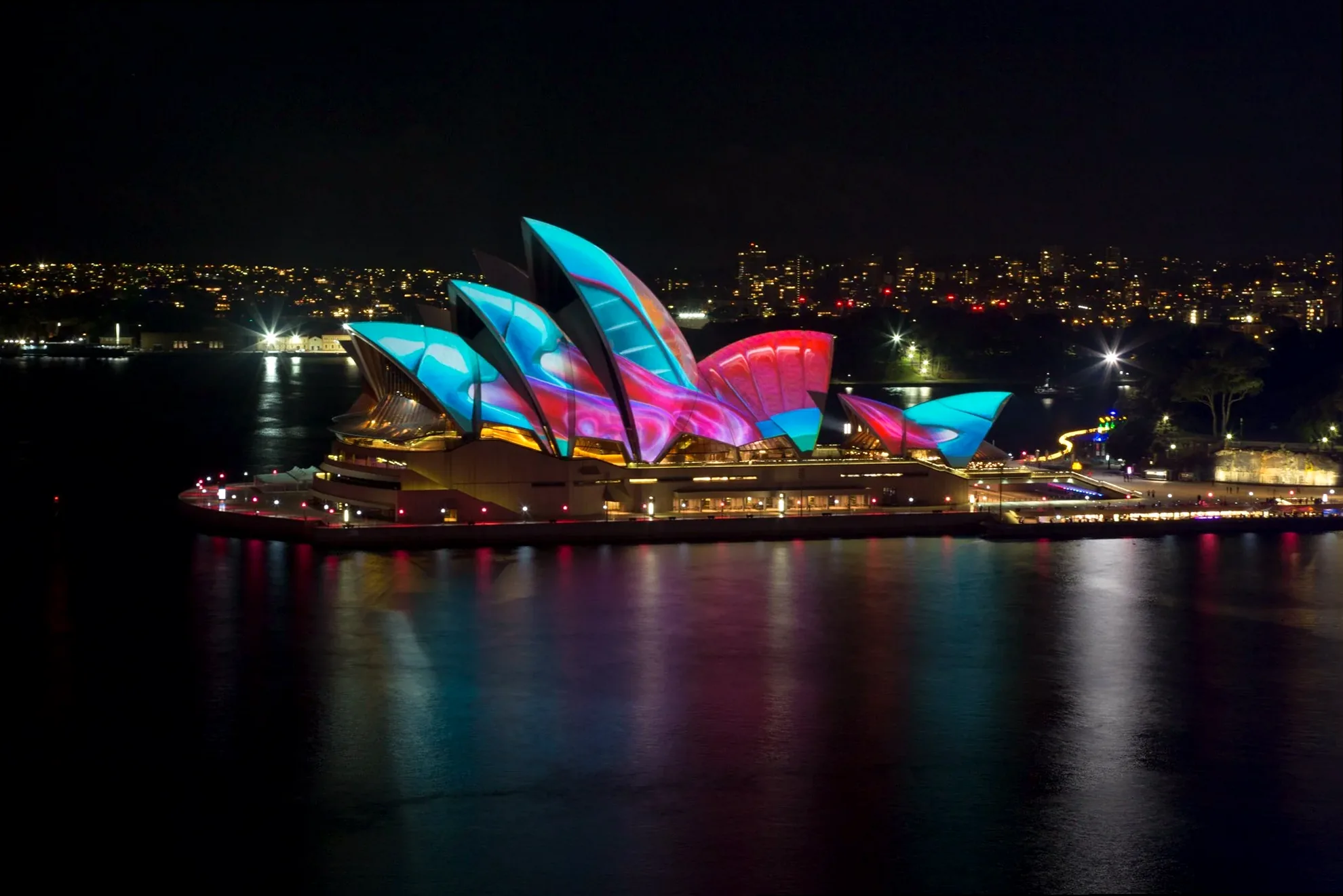 Long exposure shot of the Sydney Opera House sails glowing with intricate patterns, reflected in the water.