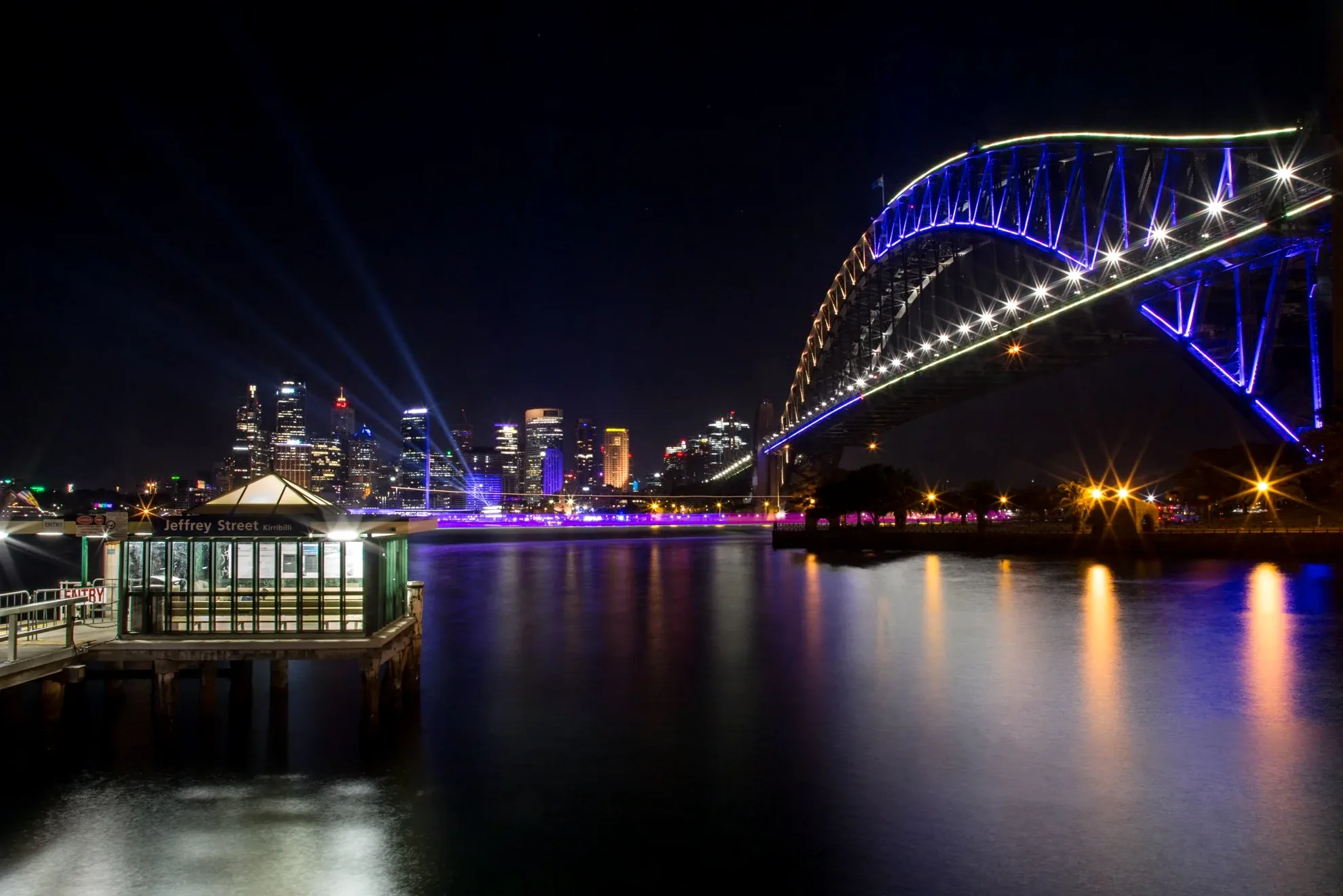 Long exposure photography of the Sydney Harbour Bridge at night, showcasing light trails and colorful illumination.