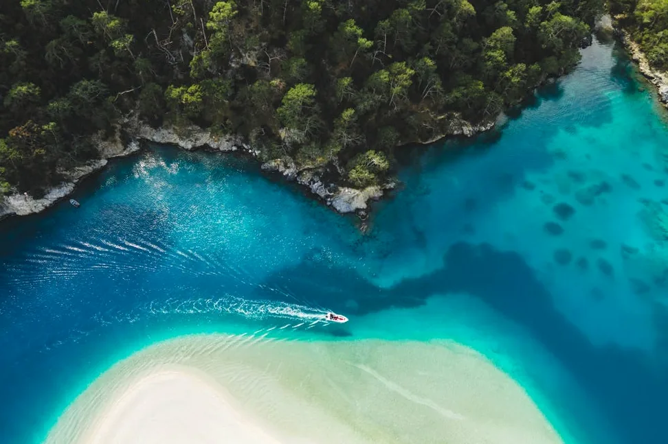Aerial drone view of Oludeniz Blue Lagoon with turquoise sea, white sand beach, and tourist boat, inspiring funny captions about paradise found or travel mishaps.